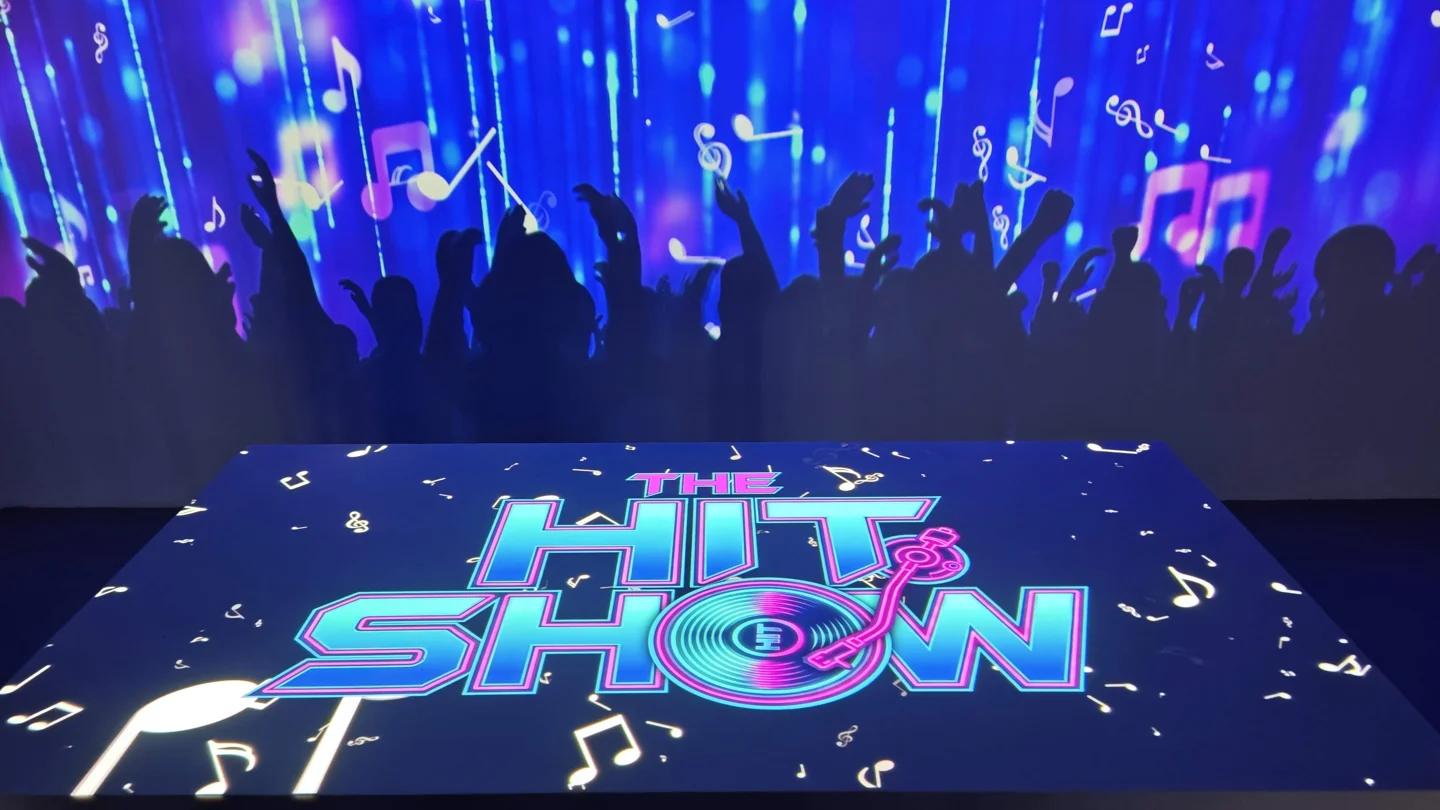Escenario inmersivo de The Hit Show con su logotipo en el suelo y siluetas de público en las paredes